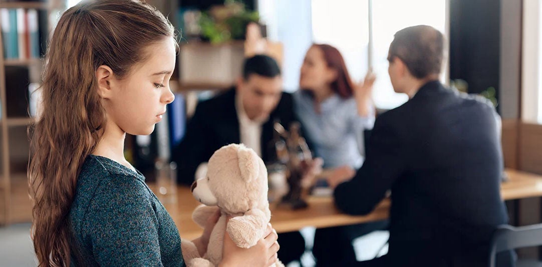 Sad girl holding teddy bear while adults argue in background, possibly a custody dispute.