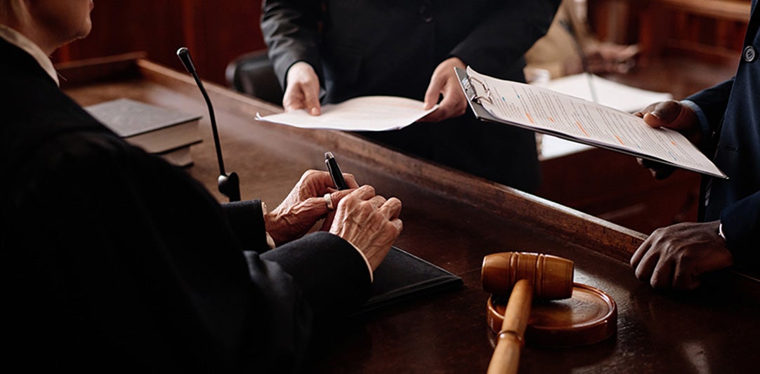 A robed judge signs papers on a wooden bench, with a gavel nearby and other people presenting documents.