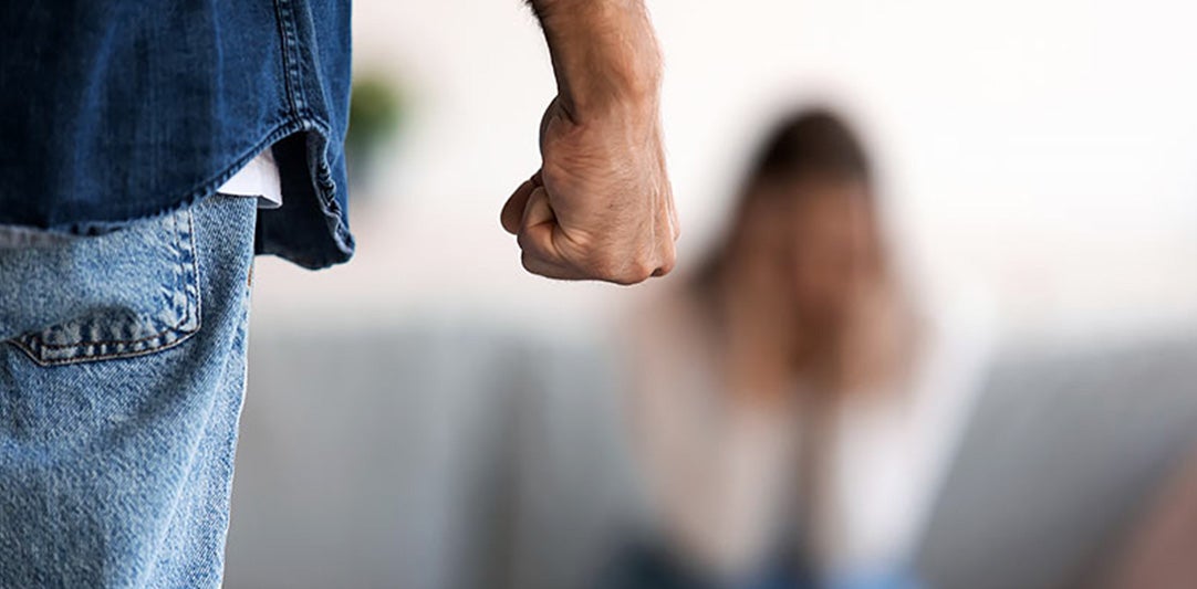 Man's clenched fist raised threateningly towards a blurred, distressed figure covering their face in the background.