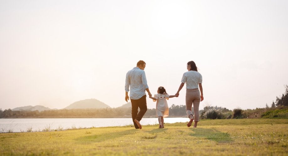 Family of three holding hands, walking away on grassy shore at sunset, with a lake and mountains.