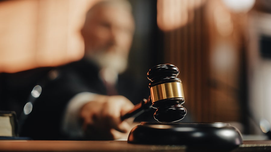 A blurred judge's hand holds a black gavel with a gold band, poised over a sounding block.